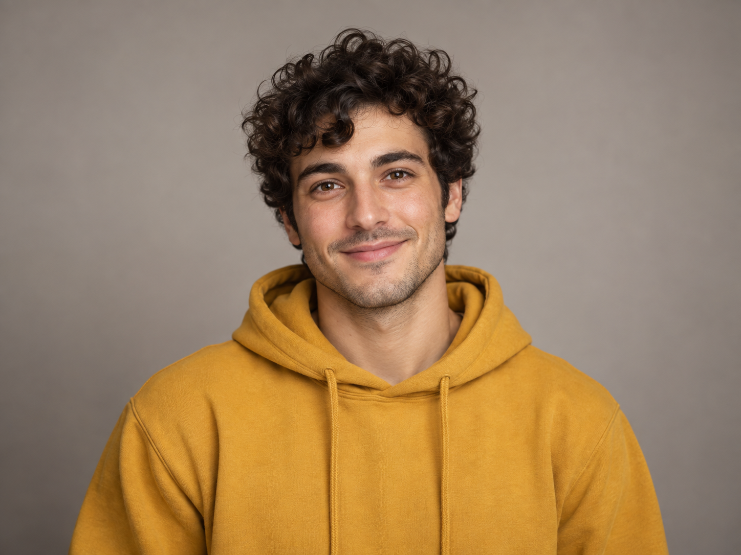 A studio portrait of a curly-haired man in a mustard hoodie